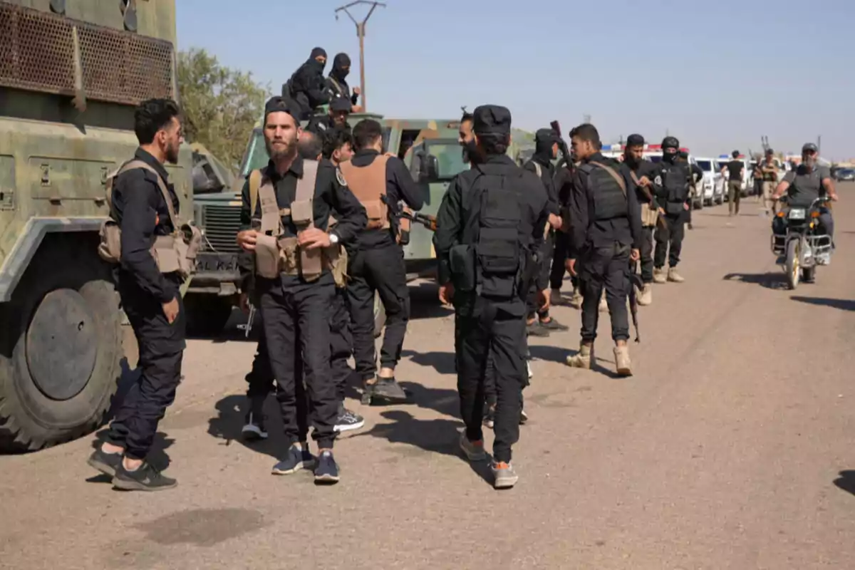 Grupo de hombres armados con uniformes oscuros y chalecos tácticos reunidos junto a vehículos militares en una carretera bajo un cielo despejado Grupo de hombres armados con uniformes oscuros y chalecos tácticos reunidos junto a vehículos militares en una carretera bajo un cielo despejado