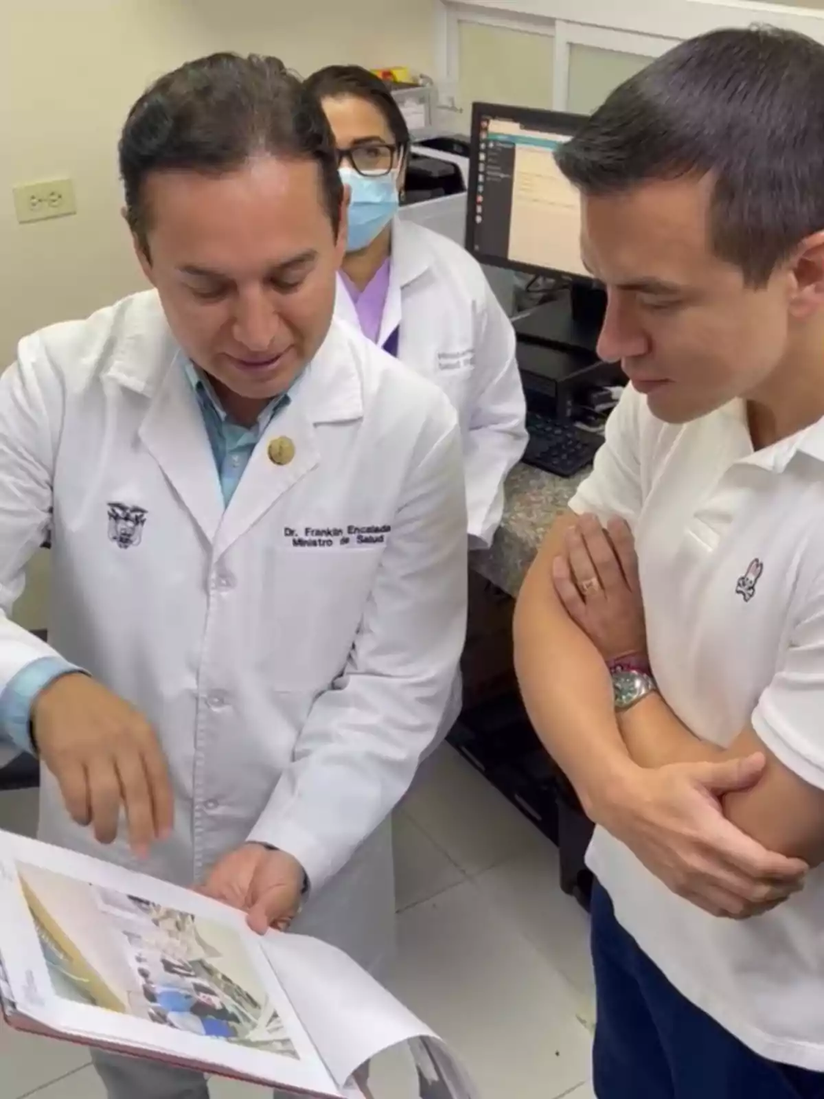Tres personas en una oficina médica observan y comentan un documento mientras una computadora está encendida al fondo