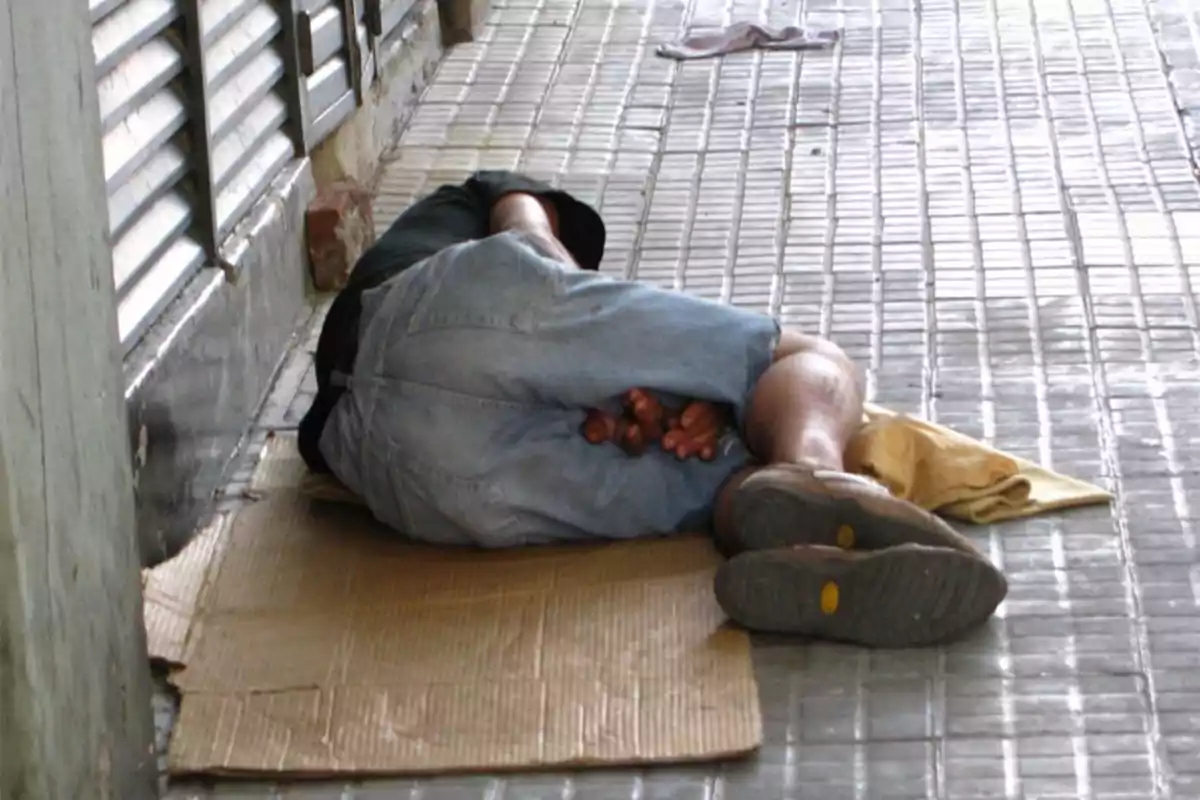 Person lying on cardboard on a sidewalk next to a wall