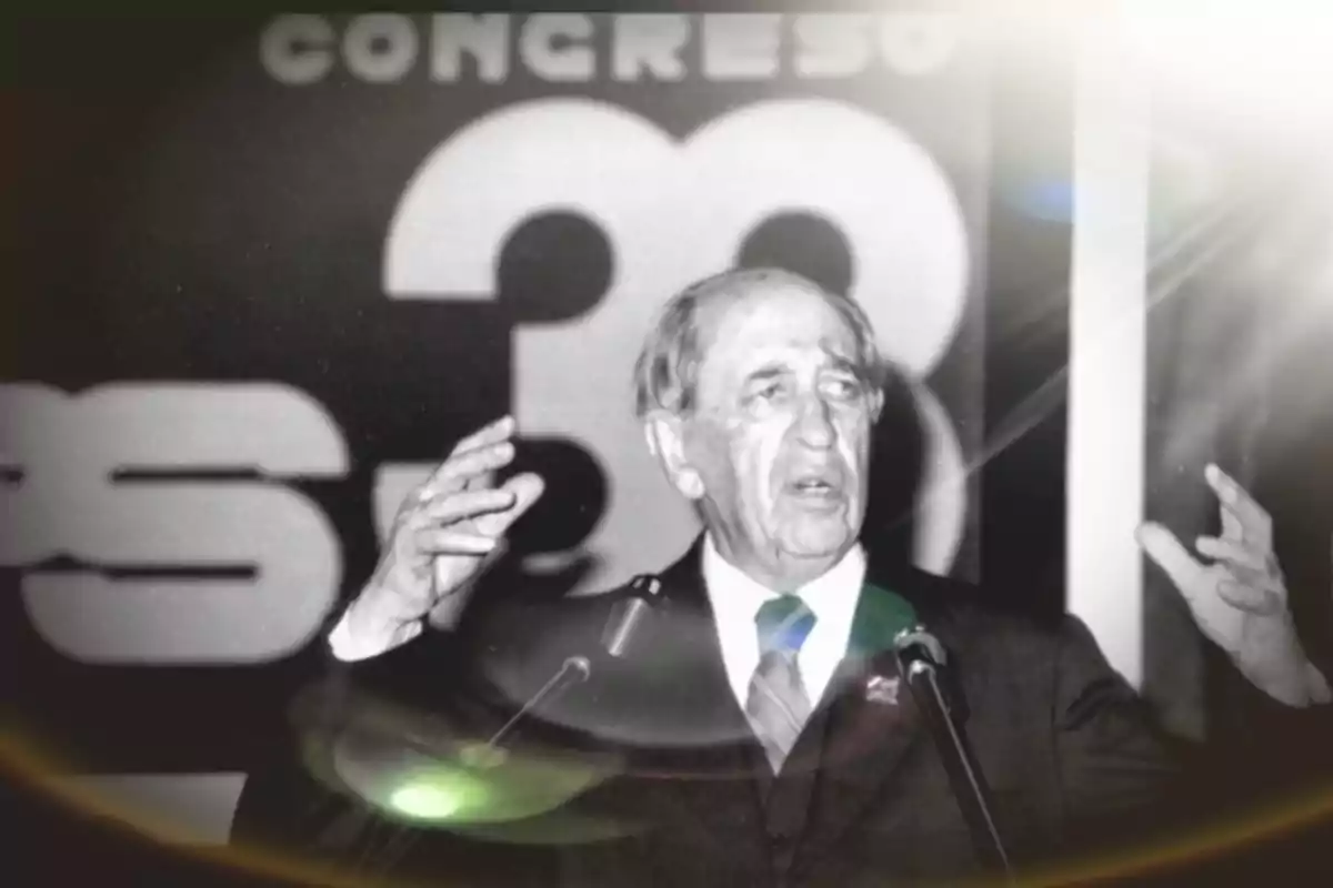 Older man in a suit speaking at a podium with his hands raised and a background with the number 38