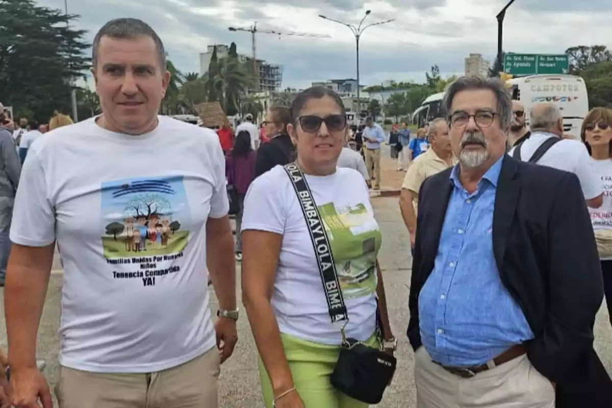 Tres personas posan para la foto en una manifestación al aire libre, dos de ellas llevan camisetas blancas con mensajes y la tercera viste saco azul y camisa celeste, al fondo se ven más participantes y un ambiente urbano. Tres personas posan para la foto en una manifestación al aire libre, dos de ellas llevan camisetas blancas con mensajes y la tercera viste saco azul y camisa celeste, al fondo se ven más participantes y un ambiente urbano.