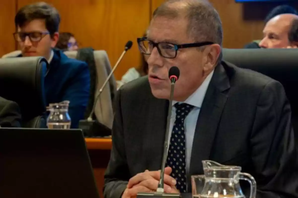 Hombre de traje y gafas hablando frente a un micrófono en una sala de reuniones con otras personas al fondo