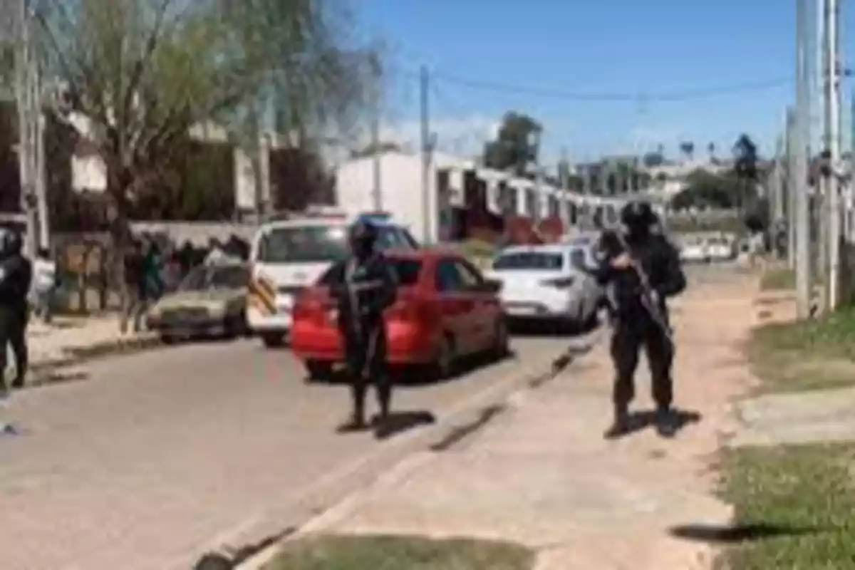Policías fuertemente armados vigilan una calle residencial donde hay varios autos estacionados y una ambulancia, mientras algunas personas observan desde la acera.