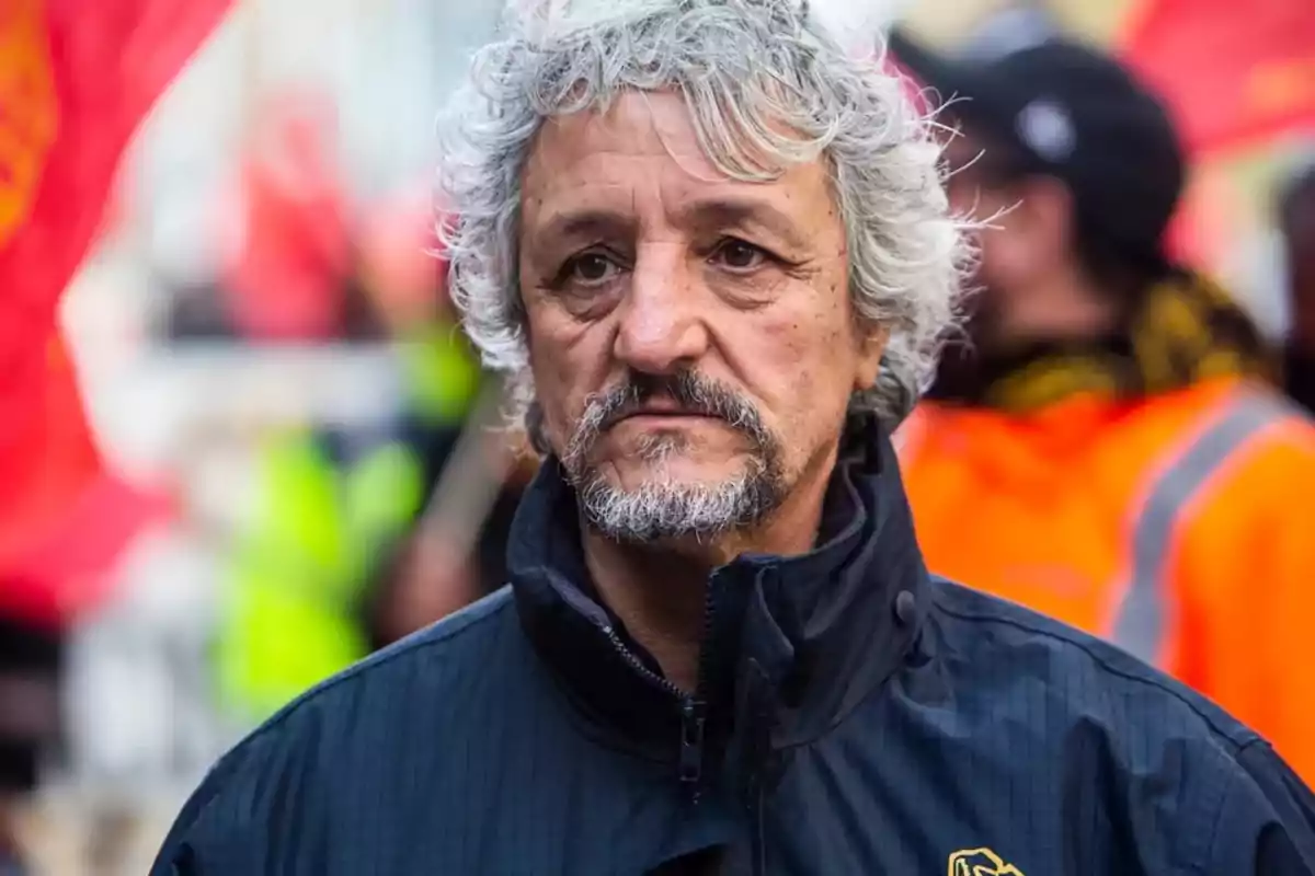 Gray-haired, bearded man with a serious expression, wearing a dark jacket, outdoors with people and blurred flags in the background Gray-haired, bearded man with a serious expression, wearing a dark jacket, outdoors with people and blurred flags in the background