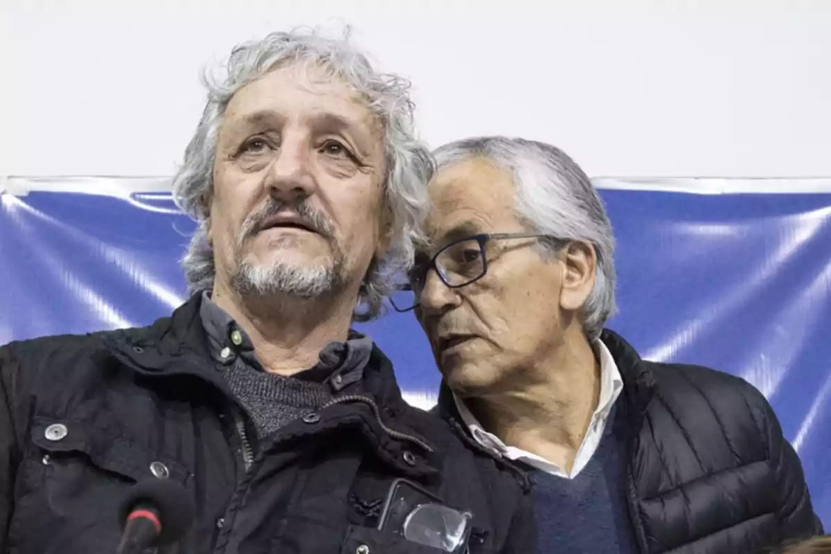 Two older men, one of them whispering in the other’s ear, both with serious expressions and a blue background. Two older men, one of them whispering in the other’s ear, both with serious expressions and a blue background.