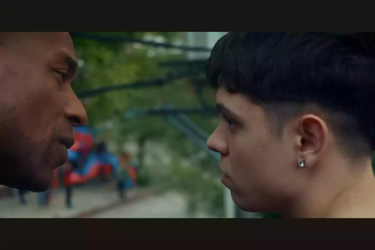 Two people stare at each other closely in an outdoor setting with trees and blurred playground equipment in the background.