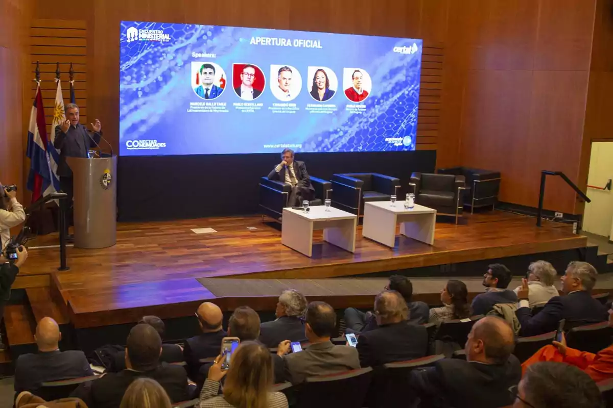 Auditorium with people attending a conference where a man is speaking at a podium and another is sitting on stage next to a large screen displaying information about the event speakers. Auditorium with people attending a conference where a man is speaking at a podium and another is sitting on stage next to a large screen displaying information about the event speakers.