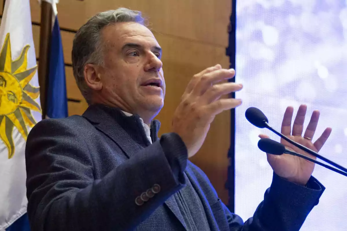 Gray-haired man speaking in public with expressive gestures, in front of microphones and a Uruguayan flag in the background