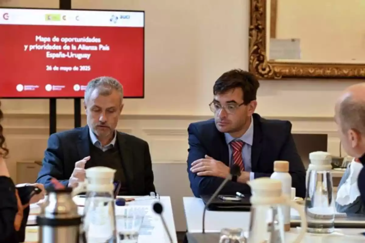 Reunión de varias personas en una sala de conferencias con una pantalla al fondo que muestra información sobre una alianza entre España y Uruguay Reunión de varias personas en una sala de conferencias con una pantalla al fondo que muestra información sobre una alianza entre España y Uruguay