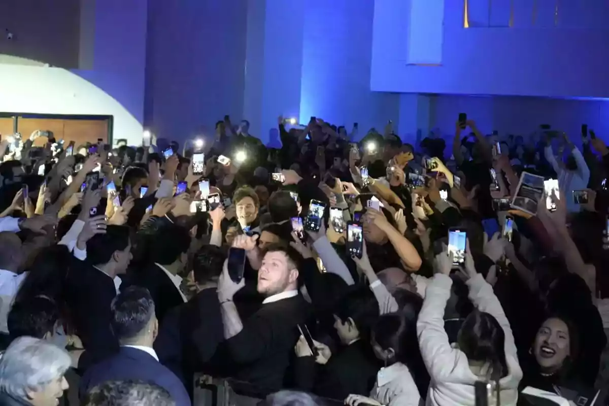 Multitud de personas en un evento levantando sus teléfonos móviles para tomar fotos o grabar videos mientras una figura central avanza entre la multitud Multitud de personas en un evento levantando sus teléfonos móviles para tomar fotos o grabar videos mientras una figura central avanza entre la multitud