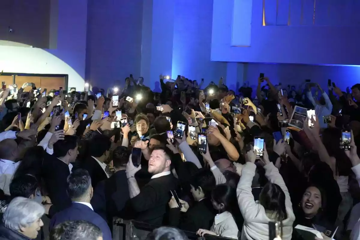 Una multitud de personas rodea a un hombre mientras todos levantan sus teléfonos móviles para tomar fotos o grabar videos en un evento bajo luces azules Una multitud de personas rodea a un hombre mientras todos levantan sus teléfonos móviles para tomar fotos o grabar videos en un evento bajo luces azules