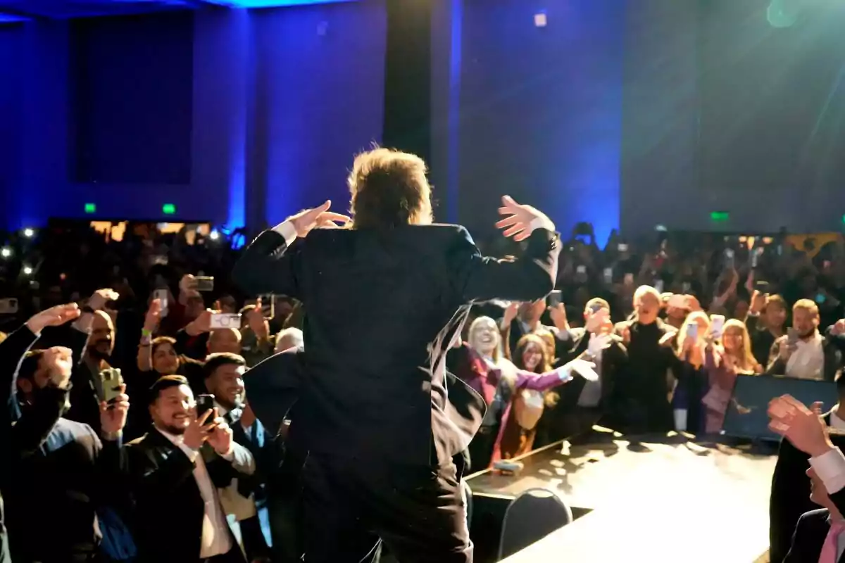 Persona de espaldas en un escenario saludando a una multitud entusiasta que toma fotos y aplaude en un auditorio iluminado con luces azules Persona de espaldas en un escenario saludando a una multitud entusiasta que toma fotos y aplaude en un auditorio iluminado con luces azules