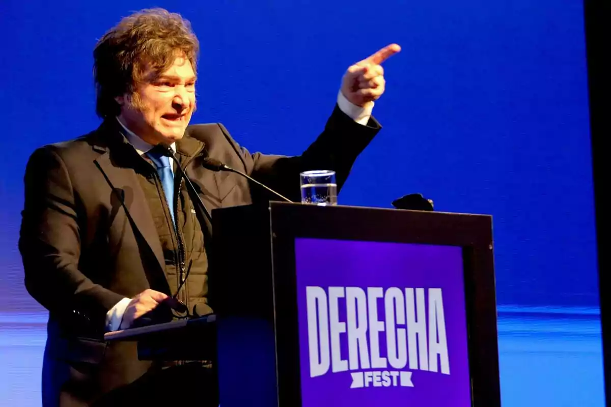 Hombre de cabello despeinado y traje oscuro hablando en un podio con el cartel DERECHA FEST sobre un fondo azul Hombre de cabello despeinado y traje oscuro hablando en un podio con el cartel DERECHA FEST sobre un fondo azul
