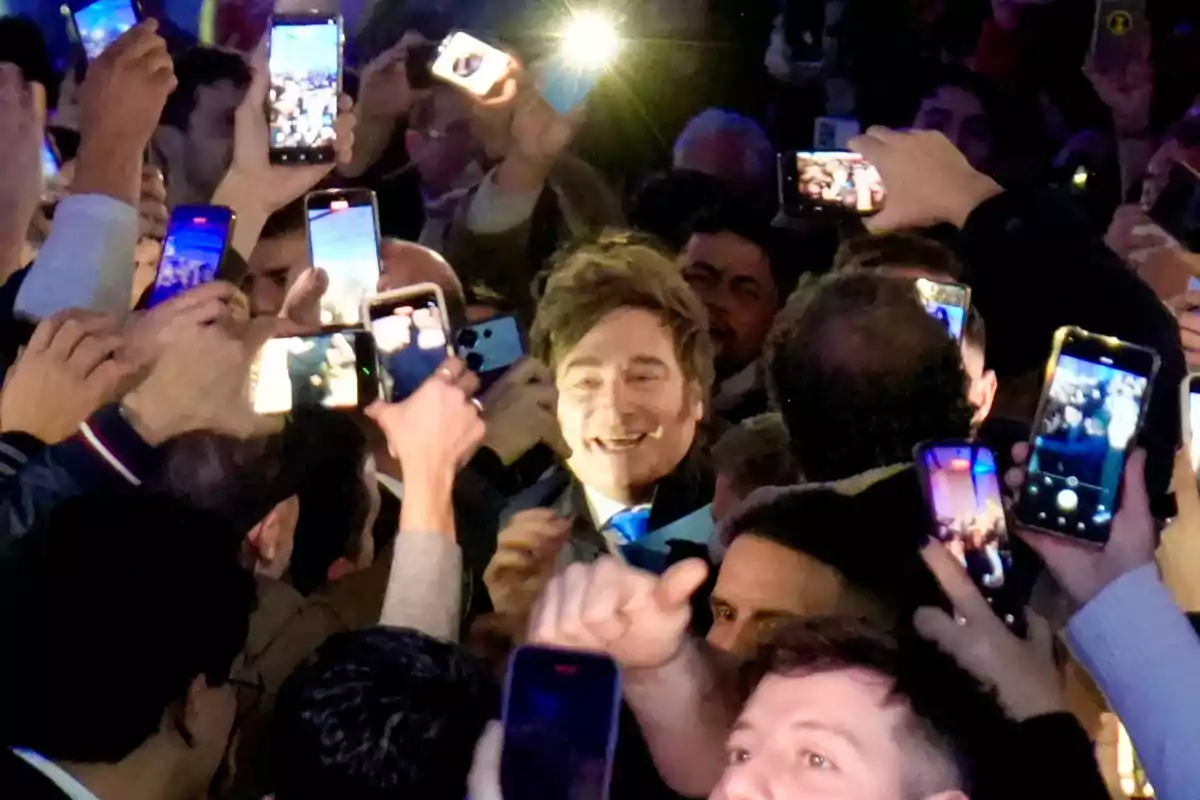 Un hombre sonriente rodeado de una multitud que lo fotografía con sus teléfonos móviles en un ambiente iluminado por flashes.