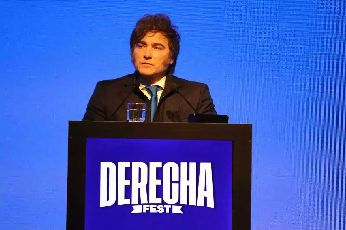 Hombre de cabello castaño y traje oscuro hablando en un podio con el texto DERECHA FEST sobre un fondo azul Hombre de cabello castaño y traje oscuro hablando en un podio con el texto DERECHA FEST sobre un fondo azul