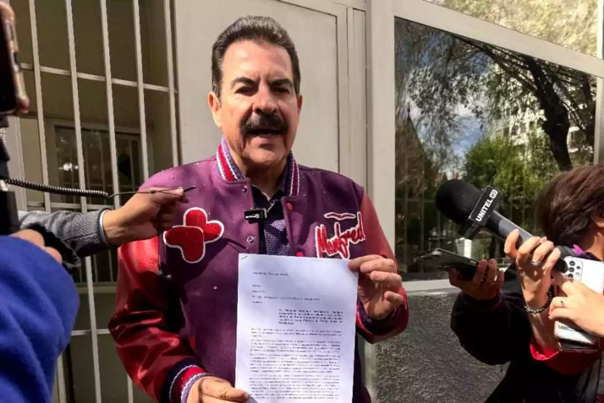 Hombre de bigote con chaqueta morada sostiene una hoja mientras es entrevistado por varios periodistas afuera de un edificio