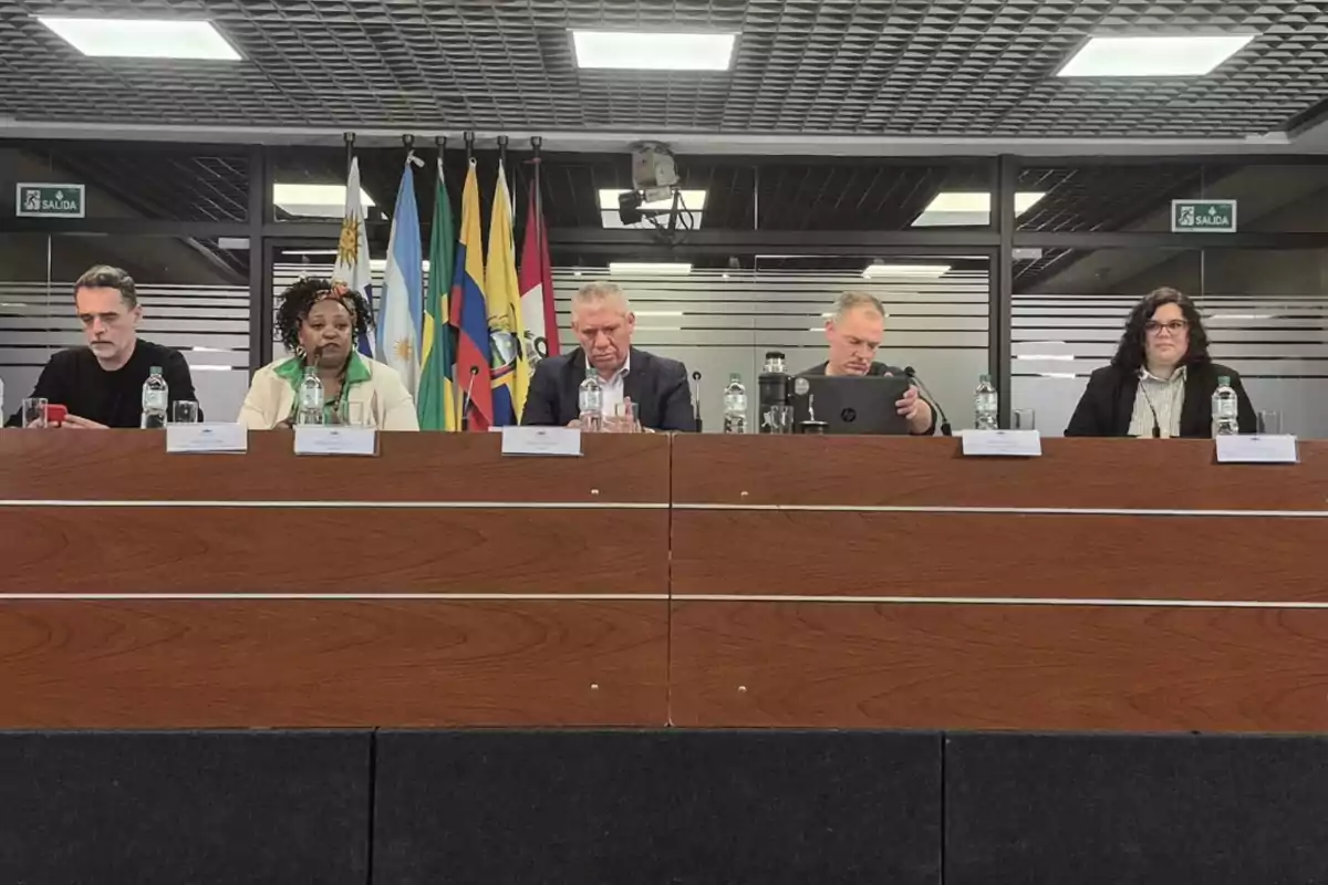 Cinco personas sentadas detrás de una mesa en una conferencia con varias banderas de países sudamericanos al fondo Cinco personas sentadas detrás de una mesa en una conferencia con varias banderas de países sudamericanos al fondo