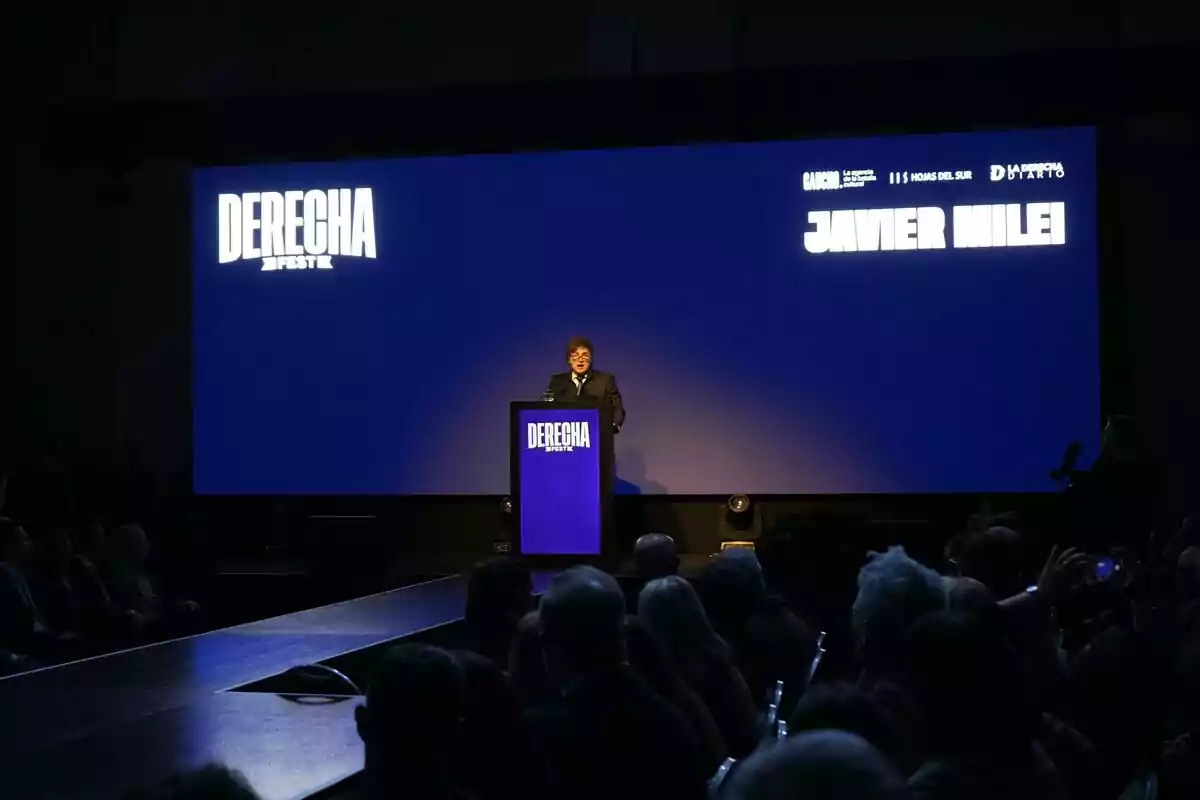 Un hombre de pie en un escenario iluminado frente a un atril azul con la palabra DERECHA FEST mientras una audiencia lo observa y una pantalla grande muestra su nombre y el evento. Un hombre de pie en un escenario iluminado frente a un atril azul con la palabra DERECHA FEST mientras una audiencia lo observa y una pantalla grande muestra su nombre y el evento.