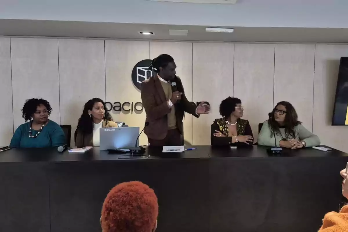 Cinco personas sentadas en una mesa de conferencia mientras una de ellas, de pie, habla por un micrófono frente a una audiencia.
