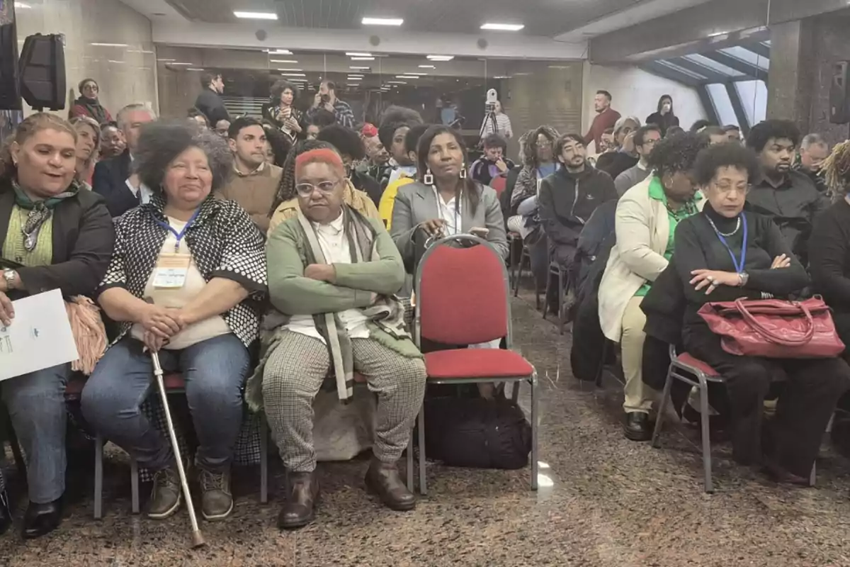 Personas sentadas en un auditorio durante un evento, algunas con gafetes y otras con bolsas, prestando atención a lo que ocurre al frente Personas sentadas en un auditorio durante un evento, algunas con gafetes y otras con bolsas, prestando atención a lo que ocurre al frente