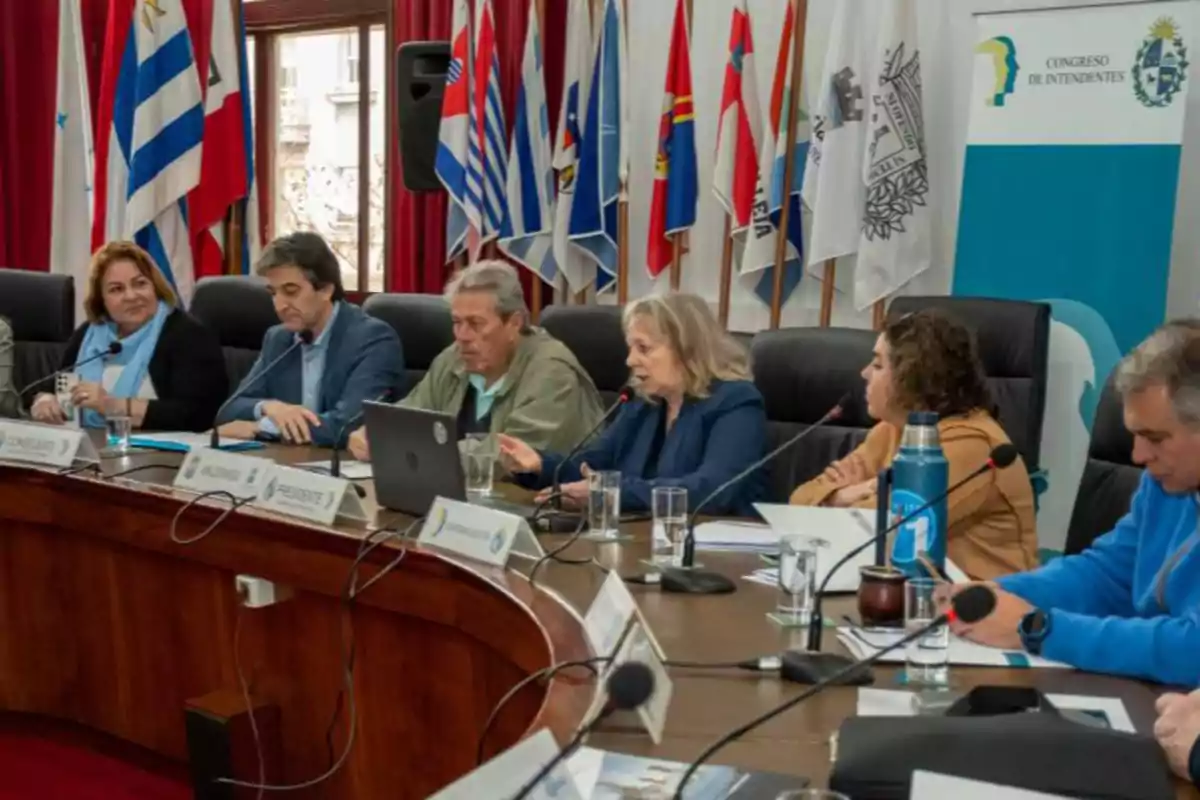 Personas sentadas en una mesa redonda participando en una reunión formal con micrófonos y documentos, al fondo hay varias banderas y un cartel que dice Congreso de Intendentes Personas sentadas en una mesa redonda participando en una reunión formal con micrófonos y documentos, al fondo hay varias banderas y un cartel que dice Congreso de Intendentes