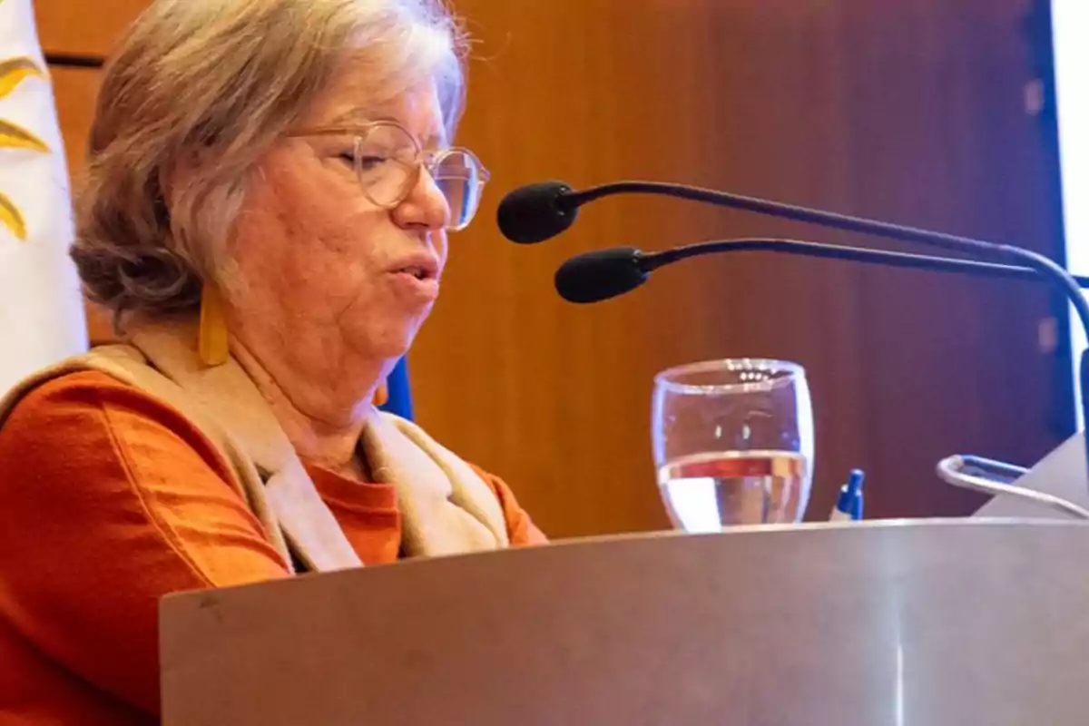 Older woman with glasses speaking in front of a microphone at a podium with a glass of water and a pen visible Older woman with glasses speaking in front of a microphone at a podium with a glass of water and a pen visible