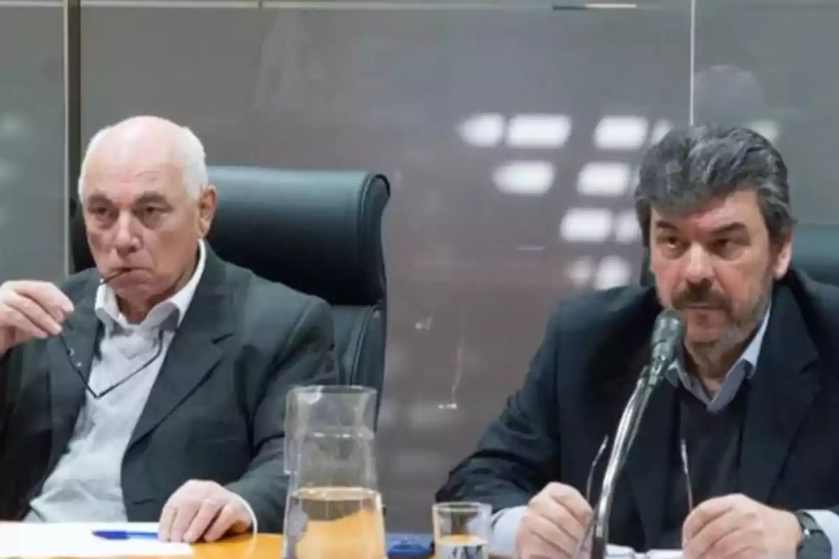 Dos hombres de traje sentados en una mesa durante una reunión o conferencia, uno de ellos sostiene unos lentes y el otro habla frente a un micrófono.