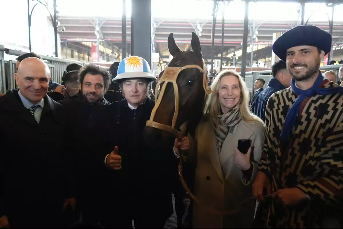 Grupo de personas posando junto a un caballo en un ambiente techado, una de ellas lleva un casco con los colores celeste y blanco y un sol amarillo, otra persona viste un poncho tradicional y boina azul