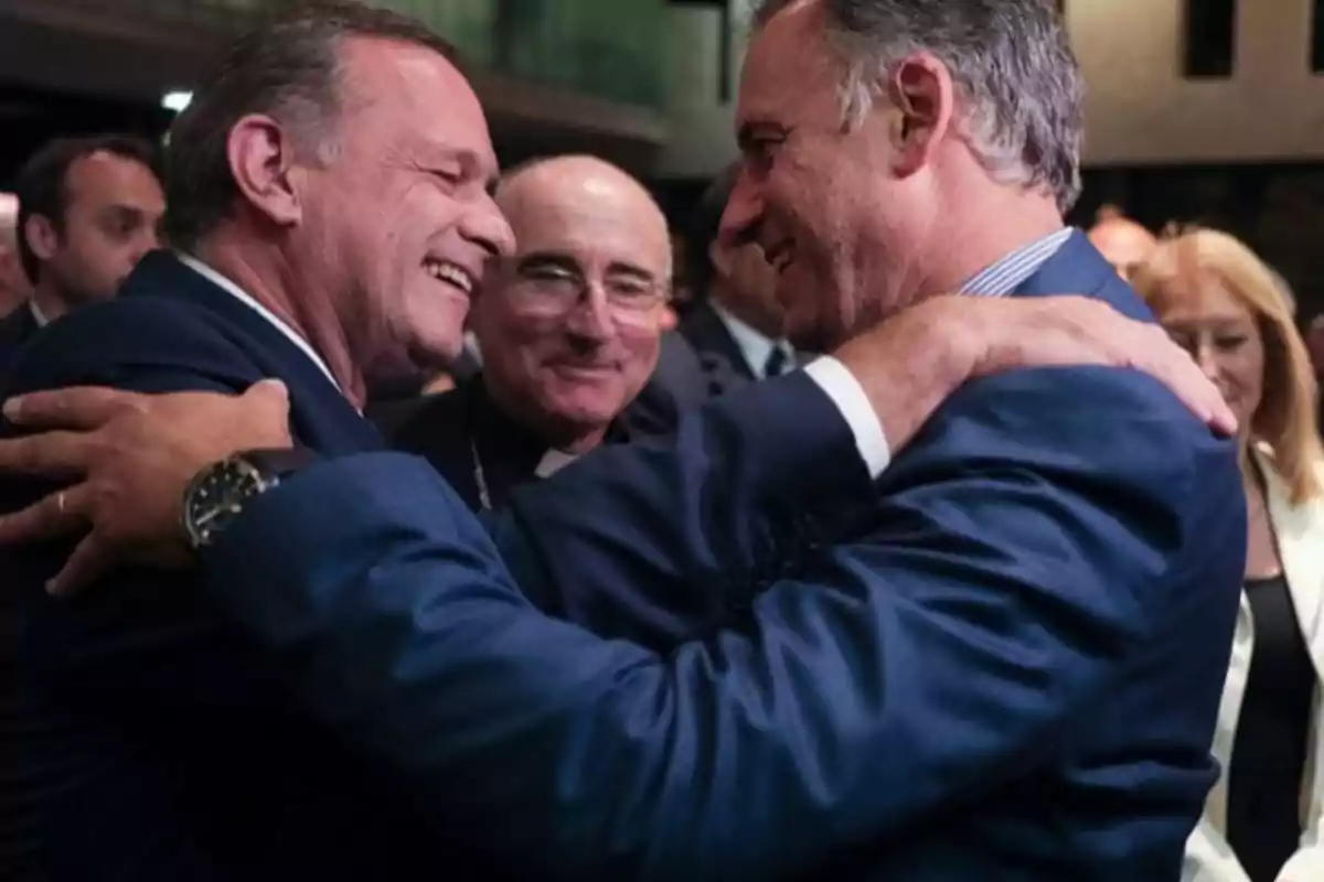 Dos hombres de traje se abrazan sonrientes en un evento social mientras otras personas observan en el fondo