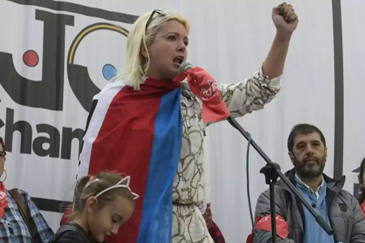 Mujer rubia con una bandera sobre los hombros hablando en un micrófono y levantando el puño en un acto público acompañada de otras personas Mujer rubia con una bandera sobre los hombros hablando en un micrófono y levantando el puño en un acto público acompañada de otras personas
