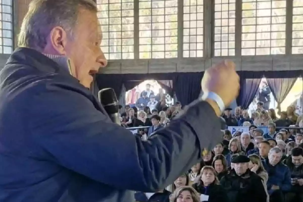 Hombre hablando con micrófono frente a una audiencia numerosa en un recinto con grandes ventanas Hombre hablando con micrófono frente a una audiencia numerosa en un recinto con grandes ventanas