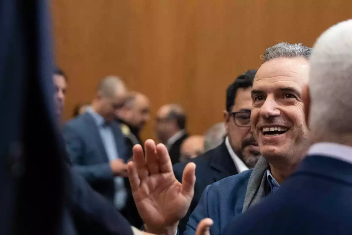 Hombre sonriente saludando a otra persona en un evento social con varias personas al fondo Hombre sonriente saludando a otra persona en un evento social con varias personas al fondo