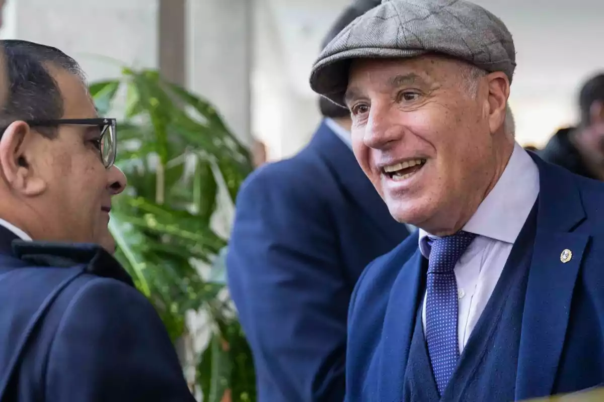 Dos hombres conversan sonrientes en un ambiente formal, uno de ellos lleva gorra y traje azul Dos hombres conversan sonrientes en un ambiente formal, uno de ellos lleva gorra y traje azul