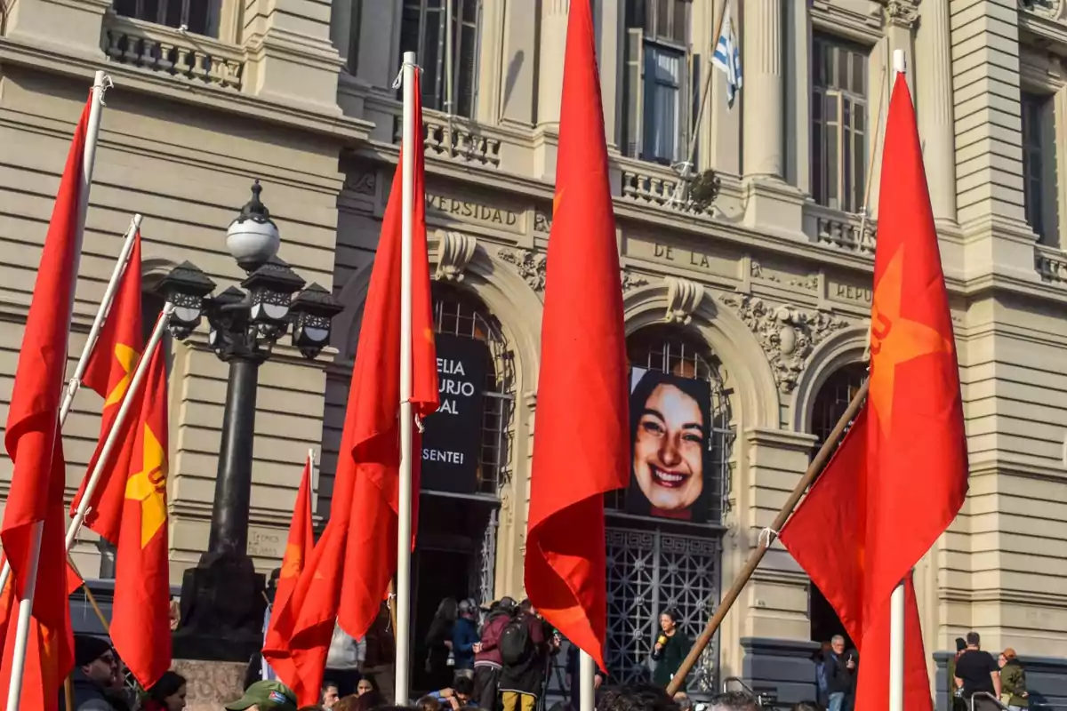 Manifestación frente a un edificio histórico con varias banderas rojas y una pancarta con el rostro de una mujer sonriente en la entrada.