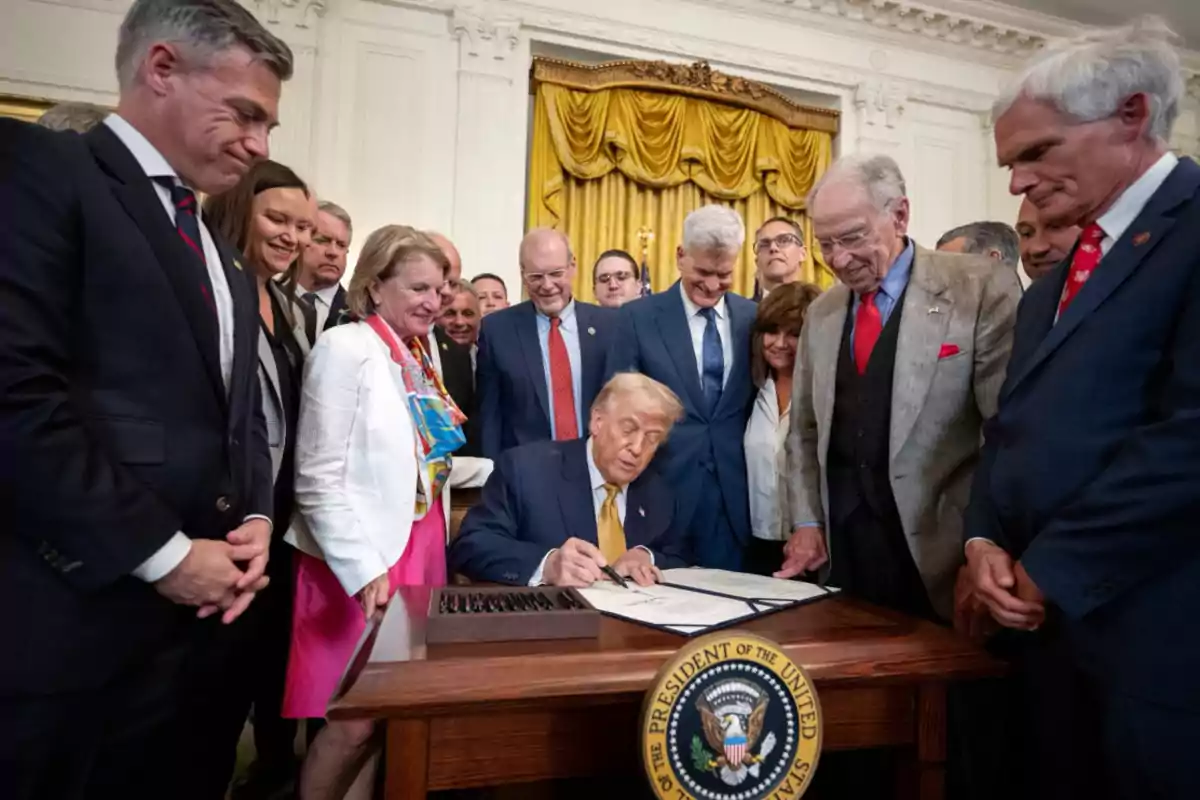 A group of people watches a man signing a document at a presidential desk in an elegant room with golden curtains.