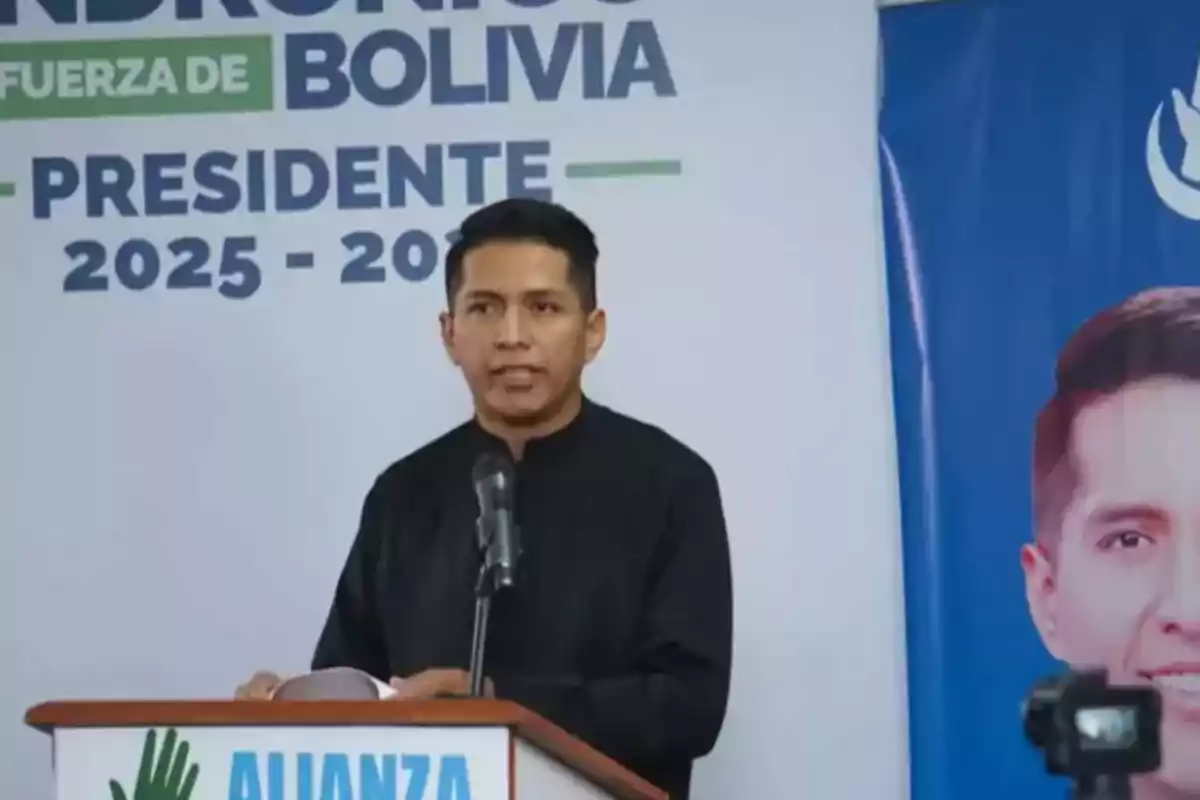 Un hombre de pie frente a un micrófono da un discurso en un podio con carteles de campaña política al fondo Un hombre de pie frente a un micrófono da un discurso en un podio con carteles de campaña política al fondo