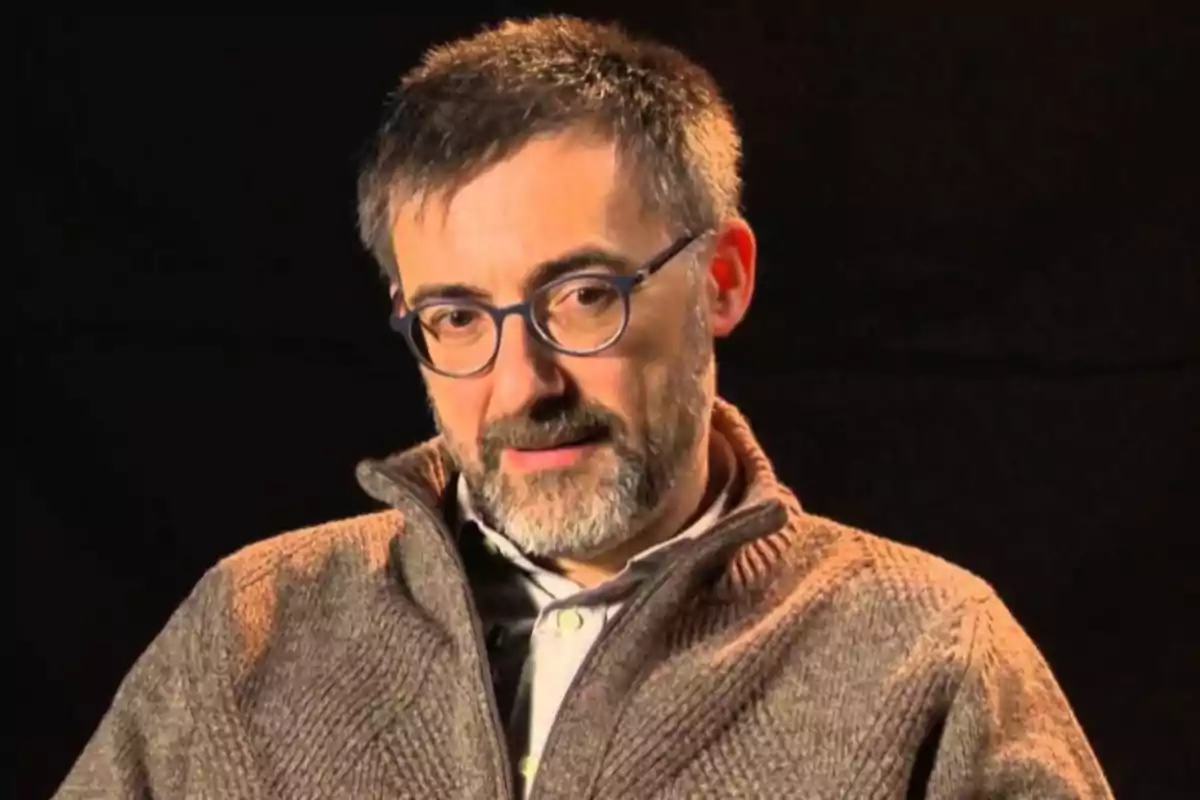 Hombre de cabello corto y barba con gafas y suéter marrón sentado frente a un fondo oscuro Hombre de cabello corto y barba con gafas y suéter marrón sentado frente a un fondo oscuro