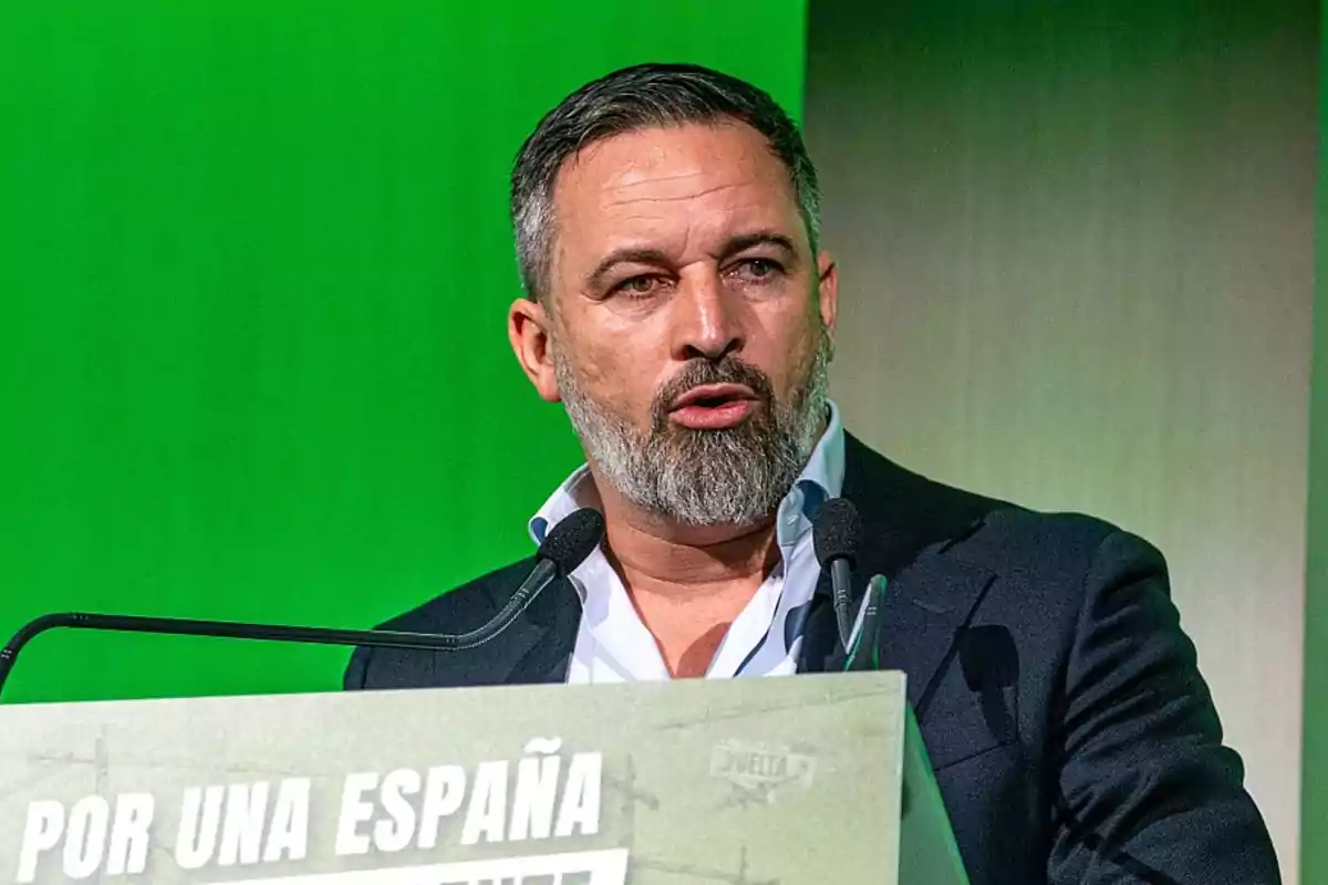 Hombre de barba y cabello corto hablando en un podio con fondo verde y un cartel que dice Por una España