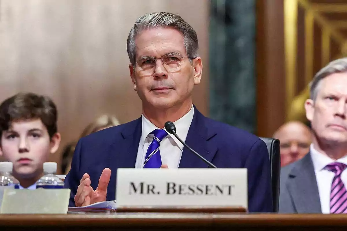 Gray-haired man with glasses seated in a formal audience with a microphone in front of him and a sign that says Mr. Bessent Gray-haired man with glasses seated in a formal audience with a microphone in front of him and a sign that says Mr. Bessent