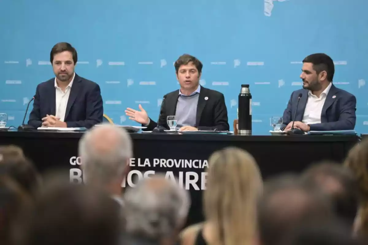Tres hombres con traje sentados en una mesa durante una conferencia de prensa frente a un público, con fondo azul y la leyenda Gobierno de la Provincia de Buenos Aires en la mesa Tres hombres con traje sentados en una mesa durante una conferencia de prensa frente a un público, con fondo azul y la leyenda Gobierno de la Provincia de Buenos Aires en la mesa