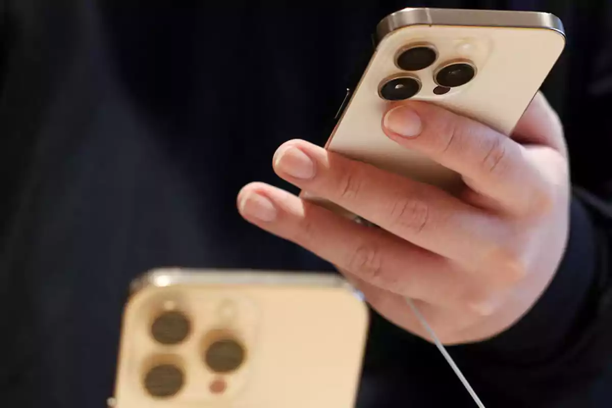 Persona sosteniendo un teléfono inteligente dorado con triple cámara trasera mientras otro dispositivo similar está sobre una mesa Persona sosteniendo un teléfono inteligente dorado con triple cámara trasera mientras otro dispositivo similar está sobre una mesa