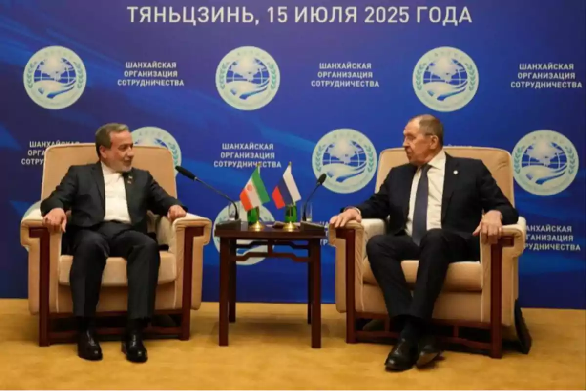 Two men in formal suits sit in armchairs conversing in front of a blue background with logos and Russian text; between them is a small table with the flags of Iran and Russia. Two men in formal suits sit in armchairs conversing in front of a blue background with logos and Russian text; between them is a small table with the flags of Iran and Russia.