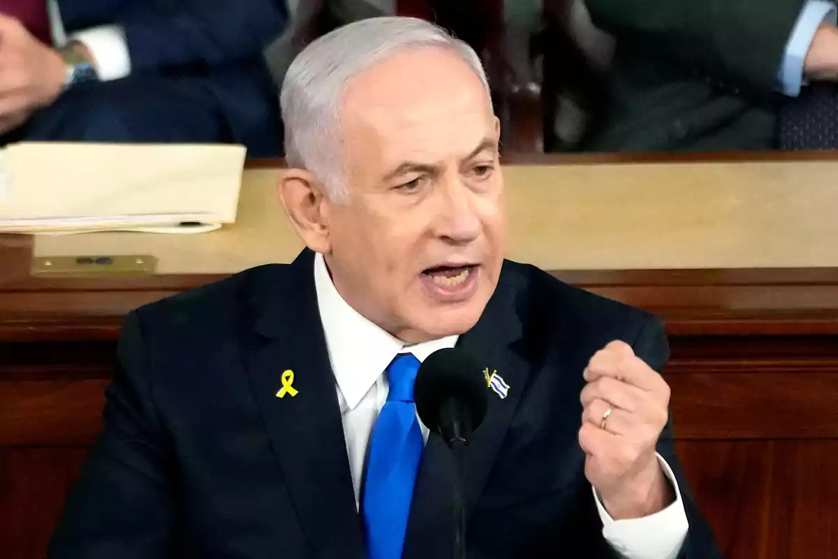Hombre de cabello canoso y traje oscuro hablando en un podio con micrófono, usando corbata azul y un listón amarillo en la solapa