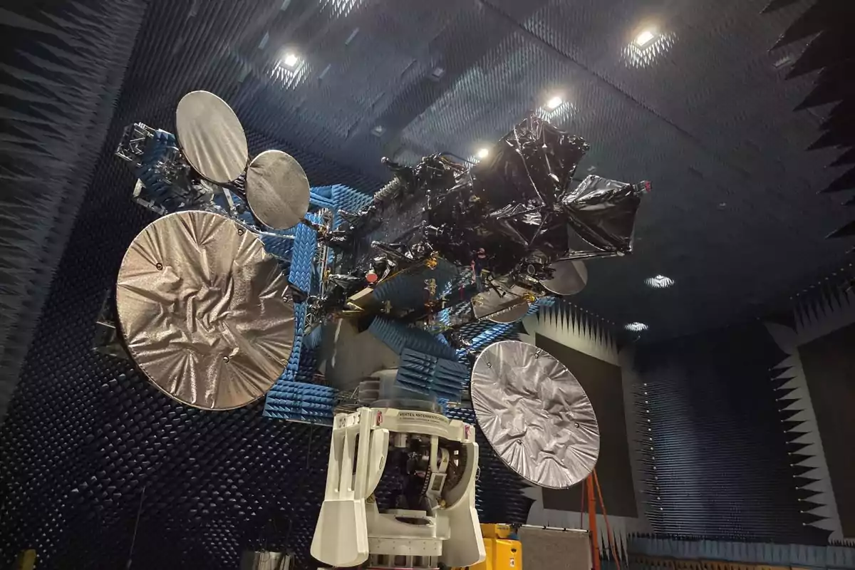 A large satellite with several circular antennas covered with reflective material is located inside an anechoic chamber with walls lined with blue and black absorbing panels. A large satellite with several circular antennas covered with reflective material is located inside an anechoic chamber with walls lined with blue and black absorbing panels.