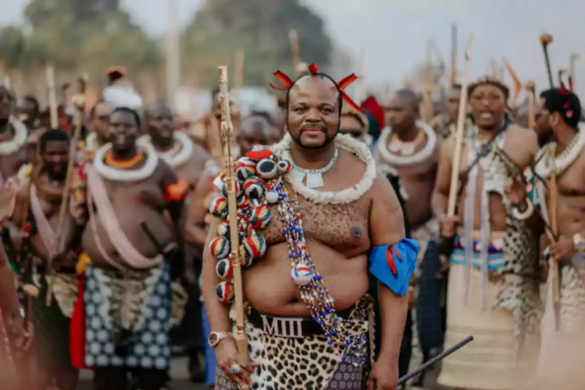 Un grupo de hombres con vestimenta tradicional africana participan en una ceremonia al aire libre, destacando uno al frente con adornos coloridos y un bastón.