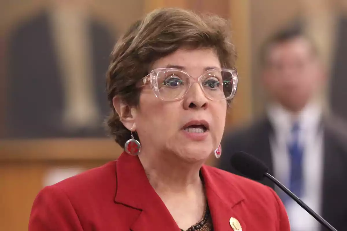 Mujer de cabello corto y gafas grandes hablando frente a un micrófono con saco rojo y aretes plateados