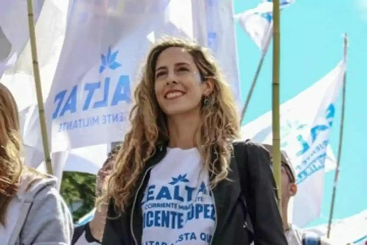 Mujer joven de cabello largo y rubio sonríe mientras participa en una manifestación con banderas blancas y celestes al fondo