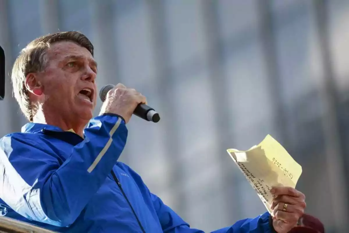 Hombre con chaqueta azul hablando por micrófono y sosteniendo una hoja de papel amarilla en la mano