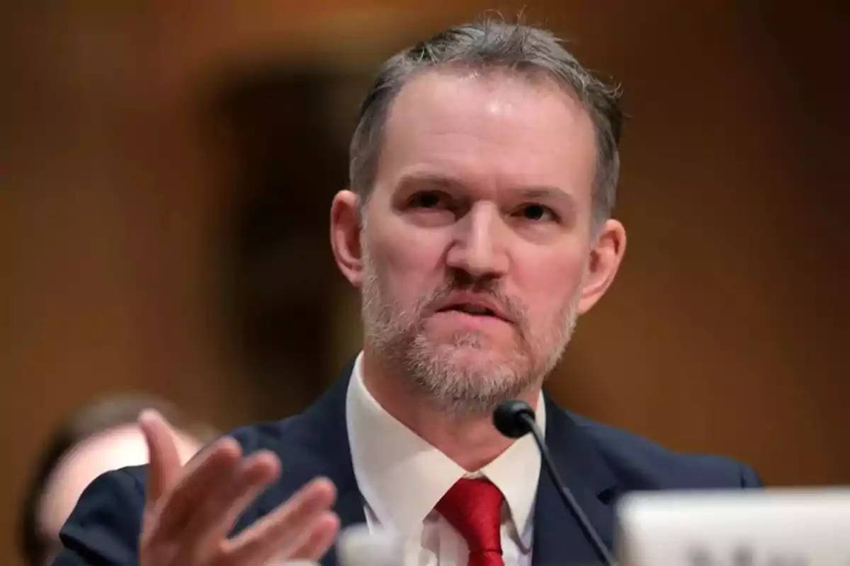 Hombre de mediana edad con barba y cabello corto, usando traje y corbata roja, hablando frente a un micrófono en un entorno formal Hombre de mediana edad con barba y cabello corto, usando traje y corbata roja, hablando frente a un micrófono en un entorno formal
