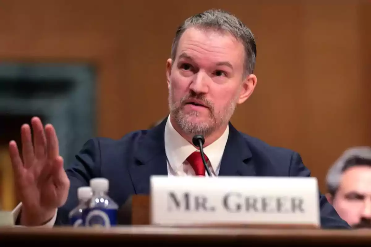 Man in a suit and tie speaking into a microphone during a hearing with a sign that says Mr. Greer in front of him Man in a suit and tie speaking into a microphone during a hearing with a sign that says Mr. Greer in front of him